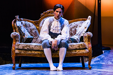 An actor in a white frilly shirt performs a scene from an opera while sitting on an ornate sofa