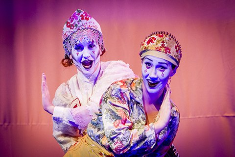 Two opera singers in expressive makeup smile as they perform on stage
