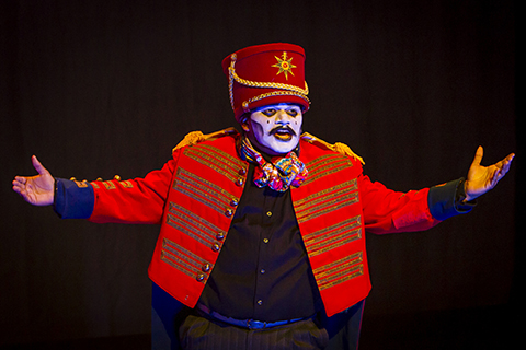 A performer in a red jacket and white makeup gestures with his arms outstretched.