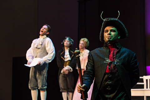 A man in a black suit and black hat stands at attention while three performers sing behind him